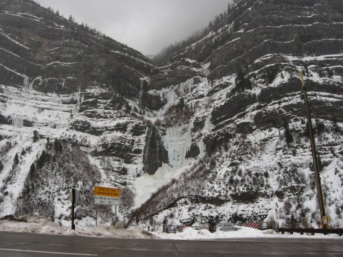 A snowy mountain landscape with a frozen waterfall cascading down rocky cliffs, under a cloudy sky.