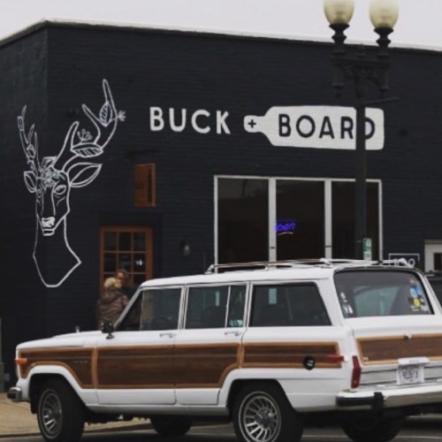 A vintage station wagon parked in front of a black building with a deer mural and the words "BUCK + BOARD."