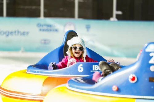 A smiling girl in a winter hat and sunglasses enjoys riding a colorful bumper car on ice.