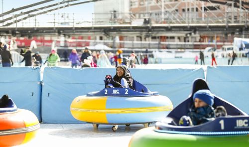 Two children enjoy bumper cars on ice, surrounded by a festive winter atmosphere and a crowd in the background.