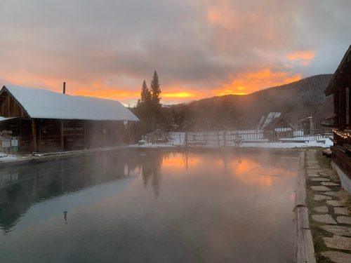 A serene hot spring at sunset, with mist rising over the water and snow-covered surroundings.