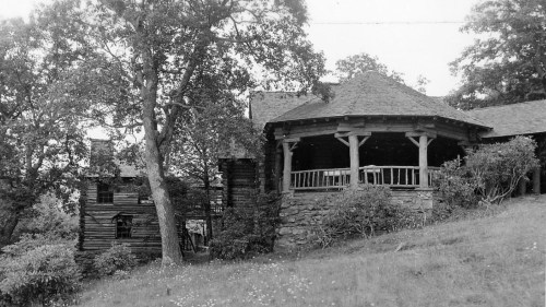 Black and white photo of a rustic house with a porch, surrounded by trees and grassy land.