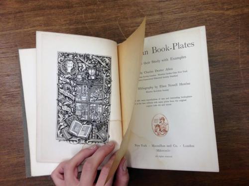 A hand holds an open book displaying an illustrated page and title about book plates.