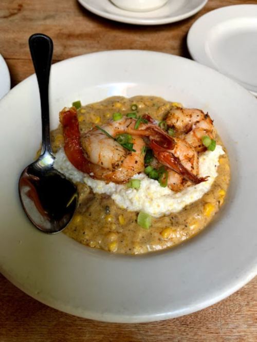 A plate of shrimp atop creamy grits with corn, garnished with green onions, served with a spoon on the side.