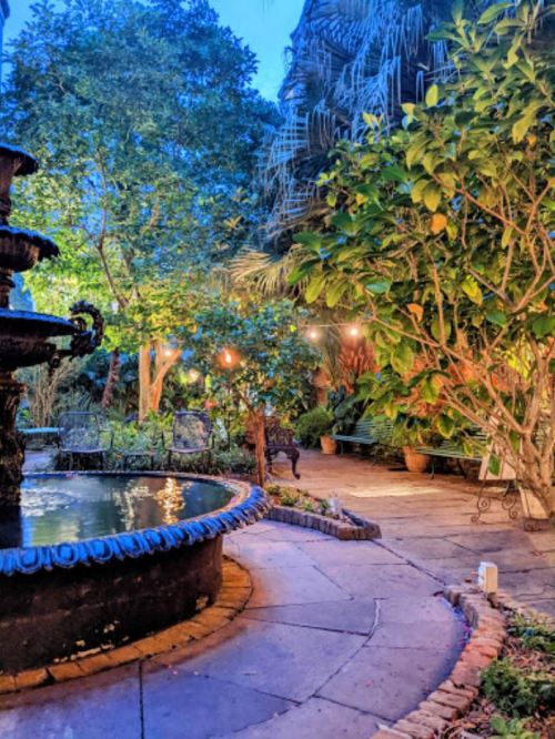 A serene garden at dusk featuring a fountain, lush greenery, and soft lighting among trees and benches.
