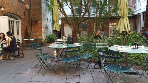 Outdoor dining area with green tables and chairs, surrounded by plants and brick walls, creating a cozy atmosphere.