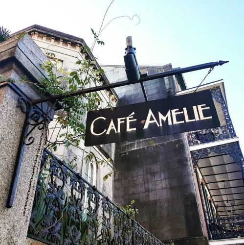 Sign for Café Amelie hanging from a wrought iron bracket, with a historic building in the background.