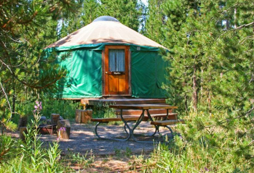 A green yurt on stilts surrounded by trees, with a picnic table and fire pit in front.