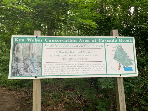 Sign for Ken Weber Conservation Area at Cascade Brook, detailing trail rules and hours, surrounded by greenery.
