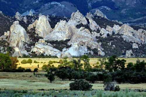 Rocky formations rise dramatically against a backdrop of mountains and a grassy field, illuminated by soft sunlight.