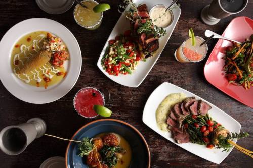 A variety of colorful dishes on a wooden table, including meats, salads, and drinks, showcasing a vibrant meal spread.