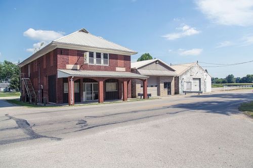 A brick building with a metal roof stands at the corner of a quiet street, with a smaller adjacent structure.