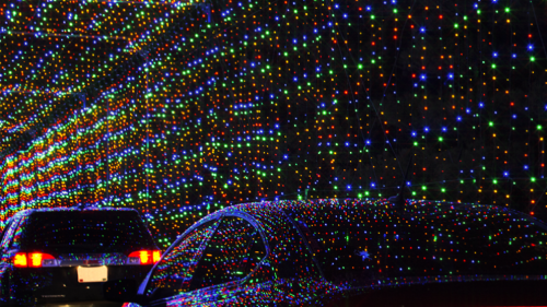 Colorful holiday lights create a vibrant tunnel effect, illuminating cars passing through.