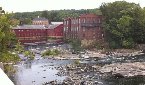 A scenic view of a river with a waterfall, surrounded by rocky terrain and historic red brick buildings in the background.