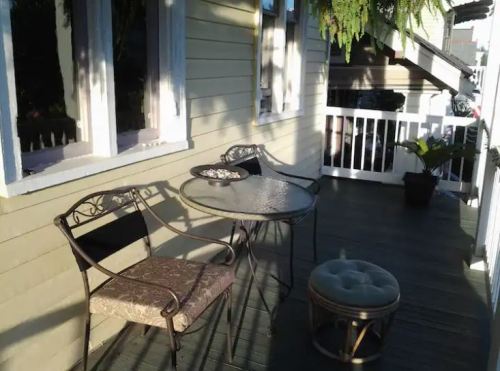 A cozy porch with a round table, two chairs, and a small ottoman, surrounded by greenery and sunlight.