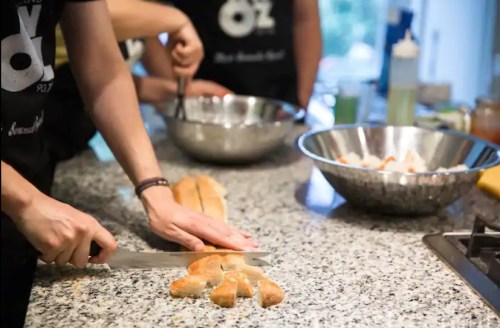 A person slices bread on a countertop while others prepare food in bowls nearby.