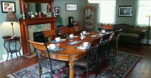A cozy dining room with a wooden table set for tea, surrounded by chairs and a decorative fireplace.