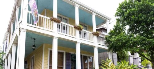 A two-story yellow house with a blue ceiling, white columns, and hanging plants on the porch.