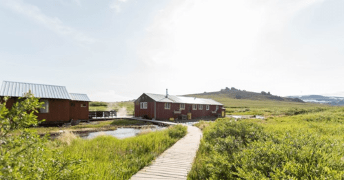 A red cabin surrounded by green grass and hills, with a wooden walkway leading to it under a bright sky.