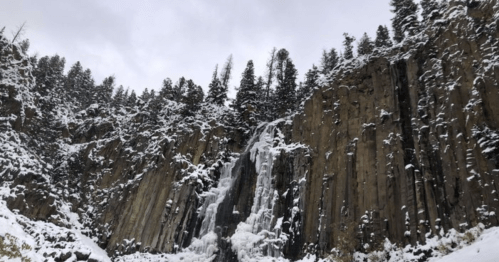 A frozen waterfall cascading down rocky cliffs, surrounded by snow-covered trees and a cloudy sky.