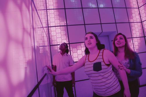 Three people interact with a colorful, illuminated wall in a modern, immersive space, expressing curiosity and excitement.