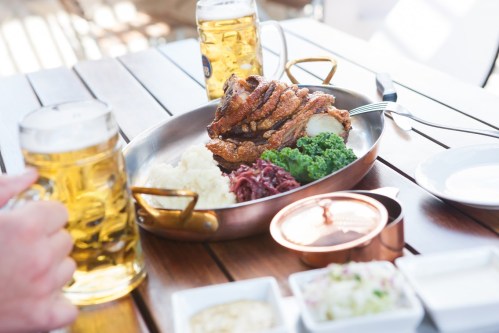 A platter of crispy pork knuckle with sides of mashed potatoes, red cabbage, and greens, alongside two beers.