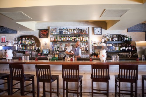 A bartender prepares drinks at a stylish bar with shelves of liquor and a warm, inviting atmosphere.