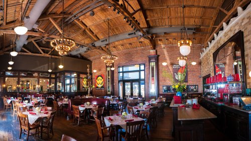 A spacious restaurant interior with wooden beams, chandeliers, and tables set for dining. Brightly decorated walls add charm.