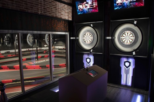 A modern dart area with two dartboards, screens above, and a view of a go-kart track through a large window.