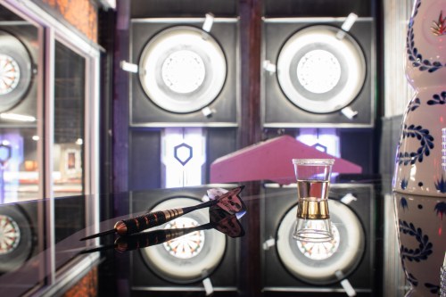 A dart and a shot glass sit on a reflective table, with dartboards visible in the background.