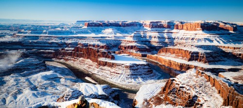 A snowy landscape of red rock formations and a winding river, showcasing a vast canyon under a clear blue sky.