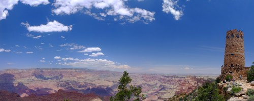 A panoramic view of the Grand Canyon with a stone watchtower against a bright blue sky and scattered clouds.