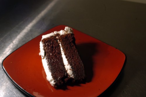 A slice of chocolate cake with white frosting on a red plate, resting on a metallic surface.
