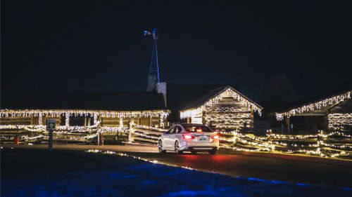 A car drives past a log cabin decorated with bright holiday lights at night.
