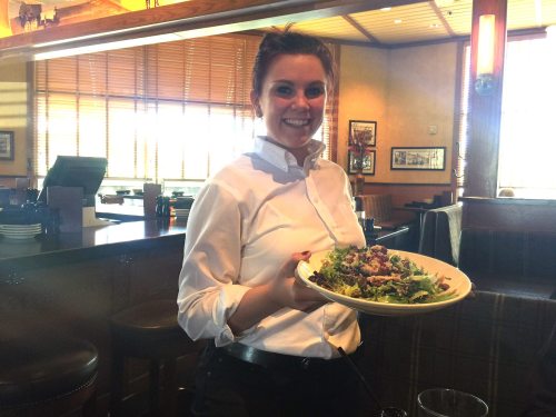 A smiling waitress in a white shirt holds a large plate of salad in a restaurant setting.