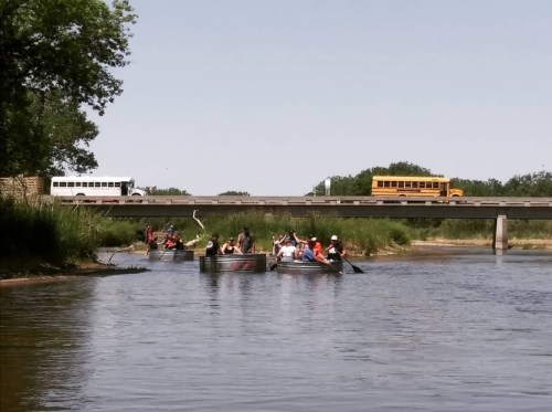 A group of people in canoes paddles on a river, with a bridge and a school bus in the background.