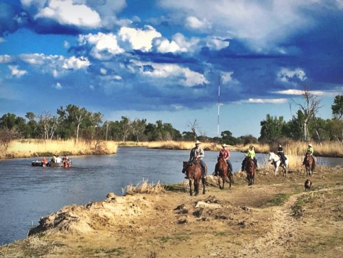 A group of horseback riders along a riverbank, with a boat on the water and a scenic sky in the background.