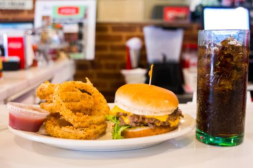 A plate with a cheeseburger, onion rings with dipping sauce, and a glass of cola on a diner table.