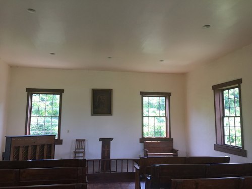 Interior of a simple, empty church with wooden benches, a podium, and large windows letting in natural light.