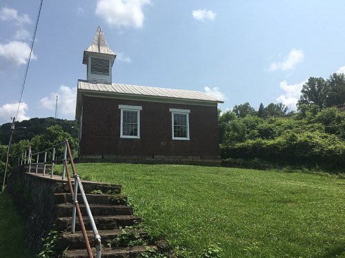 A small brick church with a steeple, set on a grassy hill under a blue sky with scattered clouds.