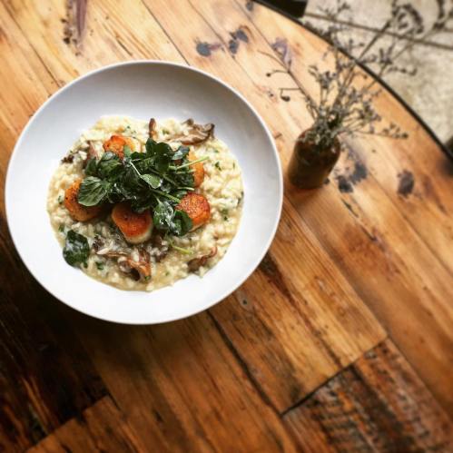 A bowl of creamy risotto topped with sautéed greens and roasted carrots, placed on a rustic wooden table.