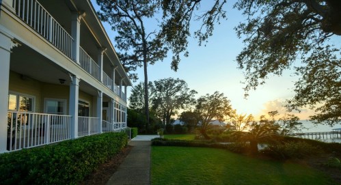 A serene sunset view from a porch, with lush greenery and a calm waterfront in the background.
