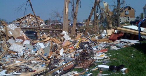 Debris and destruction from a tornado, with damaged trees and homes in the background under a clear blue sky.