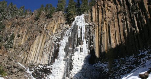 A frozen waterfall cascading down rocky cliffs, surrounded by snow-covered trees and a clear blue sky.