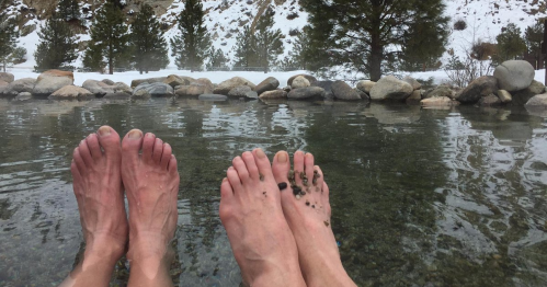 Two pairs of feet submerged in a natural hot spring, with rocks and snowy landscape in the background.