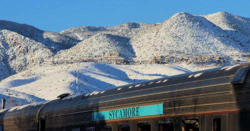 A train labeled "SYCAMORE" in the foreground, with snow-covered mountains in the background under a clear blue sky.