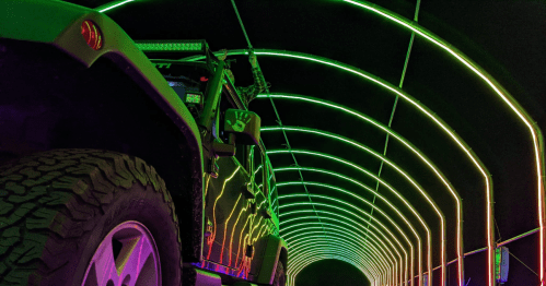A close-up of a vehicle in a brightly lit tunnel with vibrant green and blue neon lights.