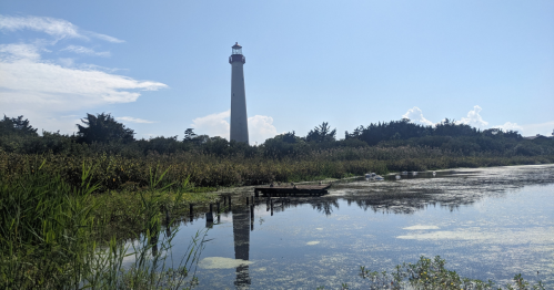 A tall lighthouse stands by a calm waterway, surrounded by lush greenery and a clear blue sky.