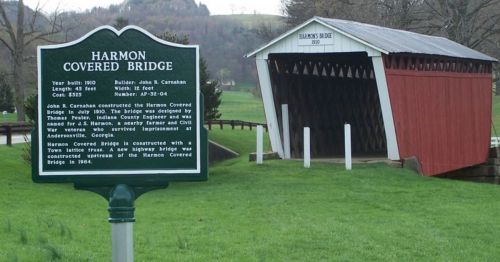 A historic sign in front of the Harmon Covered Bridge, detailing its construction and history. The bridge is red and wooden.