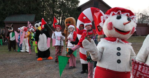 A festive line of people in holiday costumes, including a snowman, gingerbread man, and various animals, holding flags.